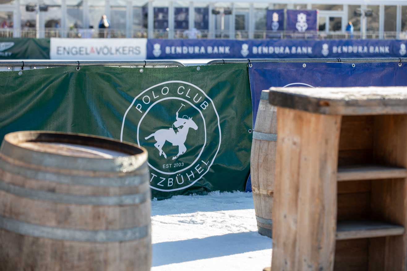 Holzfässer auf Schnee im Hintergrund grüne Bande mit weißer Aufschrift Polo Club Kitzbühel